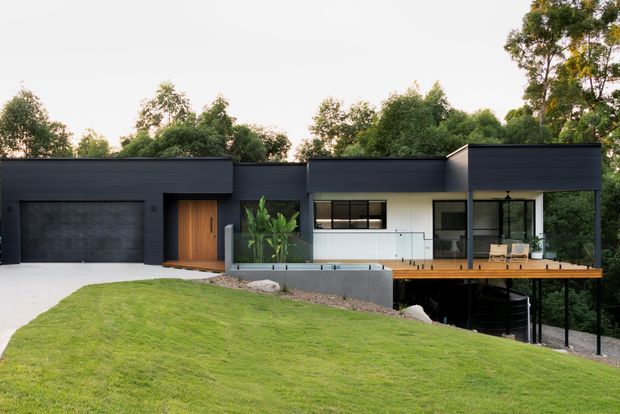 Exterior view of a contemporary single-storey house set into a sloping, landscaped site. The building has a low, flat-roofed form with dark horizontal cladding, a large integrated garage, timber entry door, extensive glazing, and a raised timber deck with glass balustrading overlooking the garden. Mature trees surround the property.