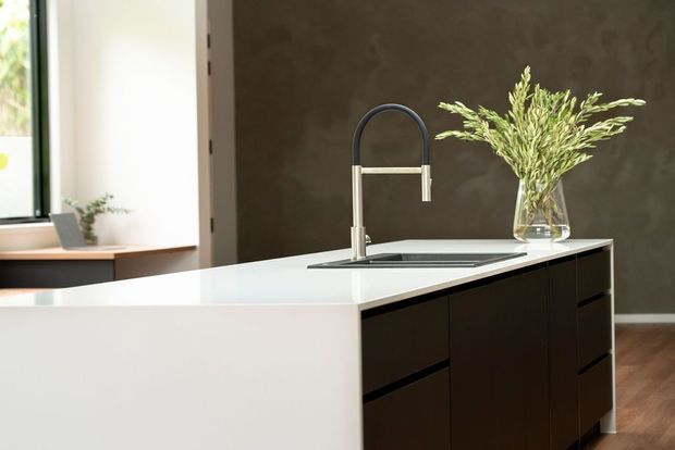 Contemporary kitchen island with a white waterfall-style benchtop, dark built-in cabinetry, integrated sink and tall curved metal tap. A large window, timber flooring and a dark textured feature wall frame the scene, with a vase of greenery on the island.