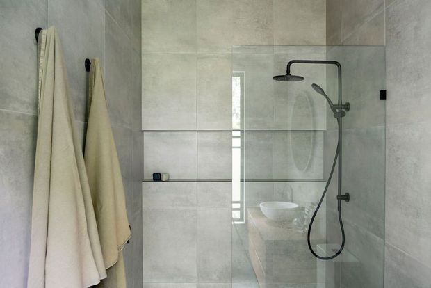 Minimal contemporary bathroom with large-format light grey wall and floor tiles, a frameless glass walk-in shower, matte black shower fixtures, and neutral towels. A wall-mounted basin is visible through the glass reflection.