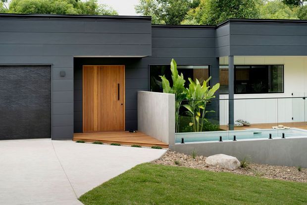 Contemporary single-storey home exterior with dark horizontal cladding, a flat roofline, timber pivot entry door, integrated garage, landscaped front garden, and a narrow pool or water feature behind a glass balustrade. The facade combines charcoal cladding with light rendered walls and warm timber detailing.