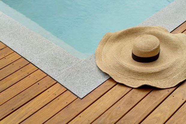 Close-up of a warm timber deck surrounding the edge of a pale blue swimming pool, with a woven straw sun hat resting on the decking. The image highlights the pool coping and timber decking materials in bright natural light.
