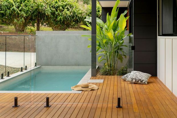 Modern outdoor pool terrace with a warm timber deck, clear frameless glass pool fencing, and a narrow turquoise swimming pool beside a dark-clad building. Tropical planting creates a lush backdrop, while a small outdoor seating area with cushions adds a relaxed resort-like character.