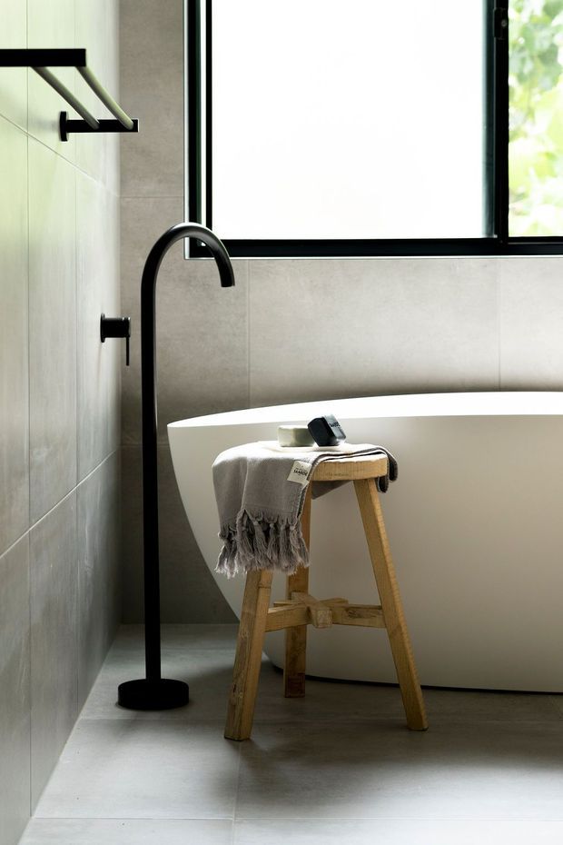 A contemporary bathroom featuring a freestanding white bathtub, large pale stone-look wall and floor tiles, matte black freestanding bath tap, black-framed window, and a simple timber stool draped with a neutral towel. The space has clean lines, abundant natural light, and a restrained neutral palette.