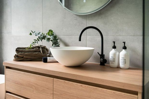A refined contemporary bathroom vanity featuring a warm timber floating cabinet, a white round vessel basin, matte black wall-mounted tapware, and neutral large-format grey wall tiles. A round mirror, folded towels, greenery, and matching soap dispensers complete the calm, spa-like composition.