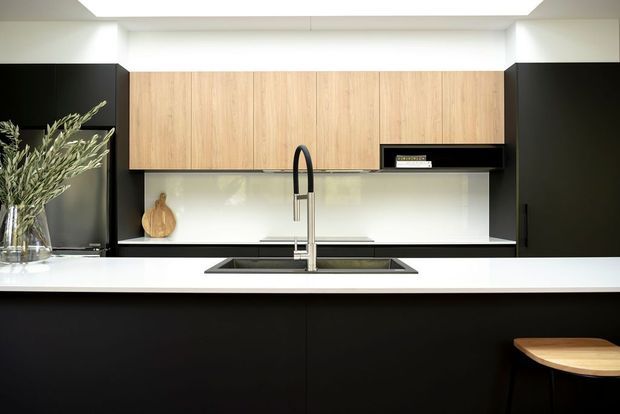 Contemporary galley-style kitchen with matte black full-height cabinetry and island base, warm timber overhead cabinets, a white benchtop, integrated black sink, tall chrome tap, and a glossy light splashback. A small timber stool and vase with foliage add subtle warmth. The concealed linear ceiling lighting creates an evenly illuminated, refined interior.
