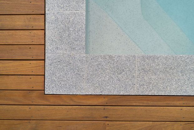 Close-up architectural detail showing a rectangular swimming pool edge set into a warm timber deck. The pool coping and surrounding border are made from speckled light-grey stone or concrete tiles, contrasting with horizontal brown timber decking. The image focuses on fixed exterior finishes and the pool surface.