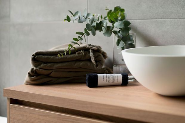 A styled bathroom vignette featuring a white vessel basin on a light timber vanity, folded taupe towels, a small eucalyptus arrangement, and a dark skincare bottle. The background is clad in large light grey concrete-look panels, creating a calm, natural and refined atmosphere.