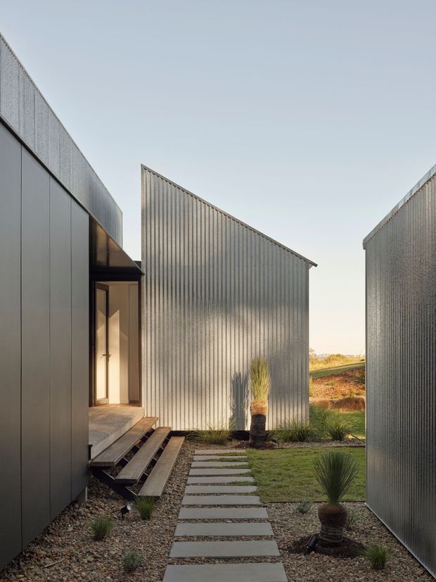 A narrow landscaped passage between several angular, corrugated metal-clad building volumes. A stepping-stone path runs through gravel and low ornamental plantings toward a raised entrance with timber steps and a glazed door. The exterior architecture has crisp geometric forms, simple pitched rooflines, and muted grey metal cladding, with open rural landscape visible beyond.