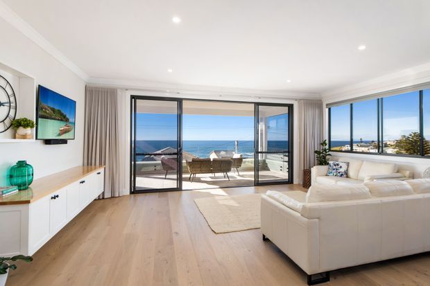 Bright, coastal-style open-plan living room with pale timber flooring, white walls and built-in media cabinetry. A large white sectional sofa faces the television, while full-height sliding glass doors and corner glazing open to a balcony with expansive ocean and coastal views.
