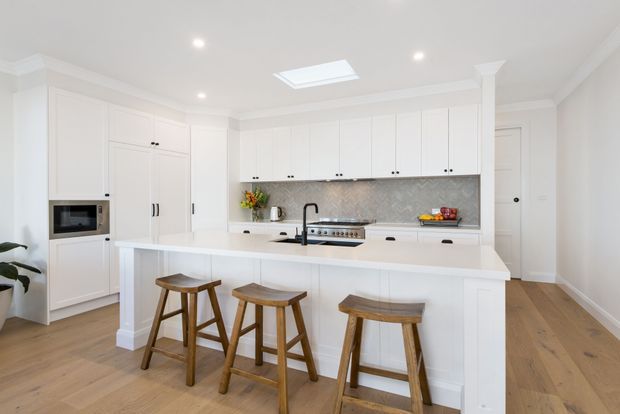 Bright, spacious white kitchen with full-height cabinetry, a large central island with seating for three, integrated microwave, freestanding-style range, dark tapware, and a grey patterned splashback. Warm timber flooring and stools add contrast, while a skylight and recessed ceiling lights provide abundant natural and artificial light.
