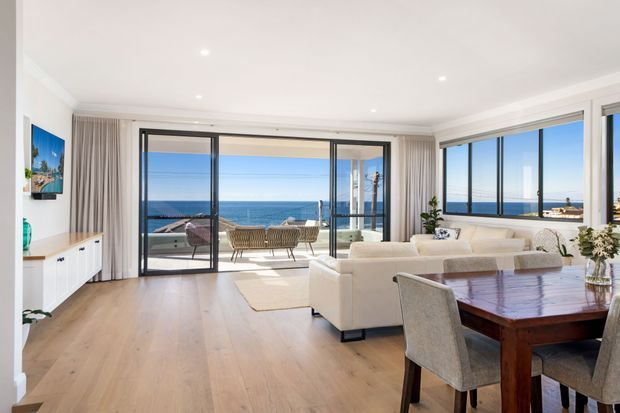 Bright contemporary open-plan living and dining room with pale timber flooring, white furnishings, expansive black-framed glazing, and direct views of the ocean. A balcony with outdoor seating is visible beyond the sliding doors, while built-in white media cabinetry and coastal artwork sit along the left wall.