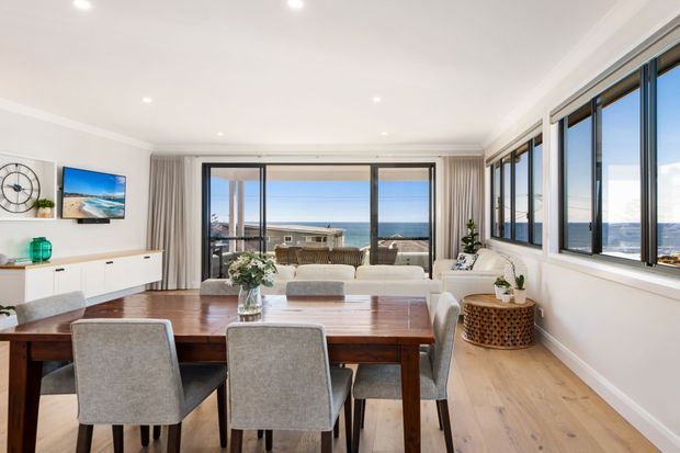 Bright open-plan dining and living area with a large timber dining table, upholstered dining chairs, pale timber flooring, white walls, recessed lighting, and extensive coastal-facing glazing. The room overlooks the ocean and includes a compact living area with a sofa, TV, and low white media console.