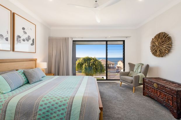 Bright, airy bedroom with a wooden bed frame and headboard, patterned aqua and grey bedding, framed botanical artwork, a grey upholstered armchair, textured curtains, plush grey carpet, and a carved wooden storage chest. Large sliding glass doors open to a balcony or exterior view, and a ceiling fan provides a relaxed coastal feel.