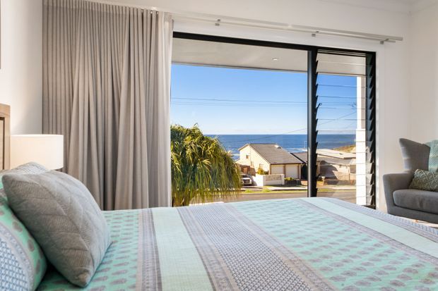 A bright coastal bedroom viewed from the bed, featuring a large ocean-facing louvred window, full-height neutral curtains, soft upholstered seating, and layered pastel patterned bedding. The window overlooks nearby homes and the blue sea.