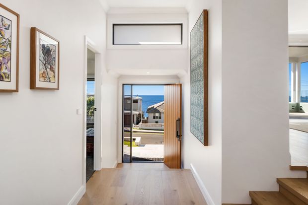Bright contemporary entry hall with white walls, pale timber flooring, framed artwork, a tall glazed front door, and a high horizontal clerestory window. The open doorway provides a glimpse toward a bedroom, while stairs and part of another bright room are visible at right. Coastal residential views are seen beyond the entrance.