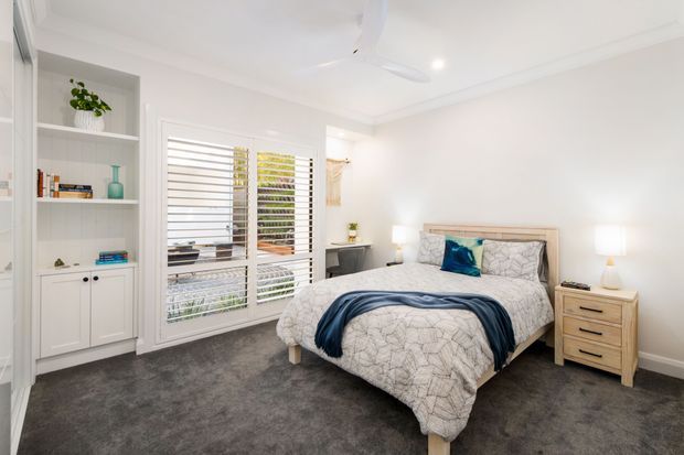 Bright, contemporary bedroom with a predominantly white palette, built-in open shelving and lower cabinetry, plantation shutters, a fabric-upholstered bed, bedside tables, and soft grey carpeting. The room has a calm, clean, lightly coastal feel with a ceiling fan and simple bedside lighting.