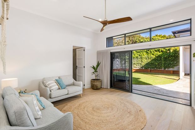 Bright contemporary living room with pale upholstered sofas, a large round natural-fibre rug, light timber flooring, white walls, and a wood-bladed ceiling fan. Full-height sliding glass doors and high clerestory windows open to a private landscaped courtyard, creating an airy indoor-outdoor connection.