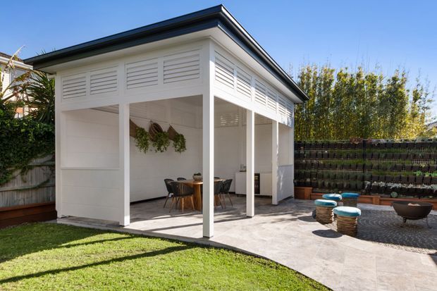A white, open-sided backyard pavilion or outdoor entertaining room with louvred upper wall panels, a dark low-pitched roof, tiled flooring, and a dining table beneath the covered structure. The space opens onto a paved patio with informal outdoor seating, raised garden beds, and a vertical planting wall.