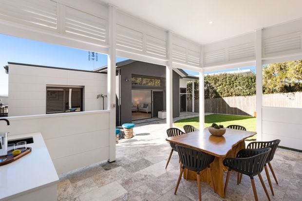 Bright covered outdoor dining and entertaining area with white louvred screening, a timber dining table and black chairs, stone-paved flooring, and views toward a landscaped courtyard and adjoining modern structures.