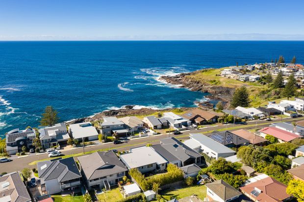 Aerial view of a coastal suburban neighbourhood overlooking a rugged rocky shoreline and deep blue ocean. The image shows detached homes with varied pitched and low-pitched roofs, streets, landscaped gardens, and a prominent headland extending along the waterfront.