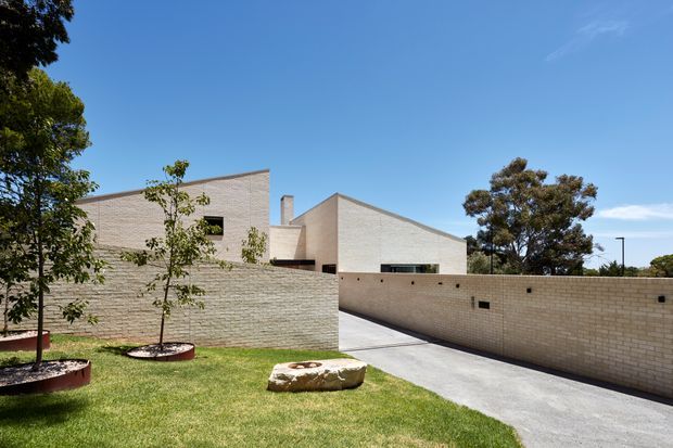 Exterior view of a contemporary low-rise brick residence with angular pitched roof forms, pale masonry walls, high-set horizontal windows, and a long brick boundary wall enclosing a sloped concrete driveway. A manicured lawn and young trees occupy the foreground under a clear blue sky.