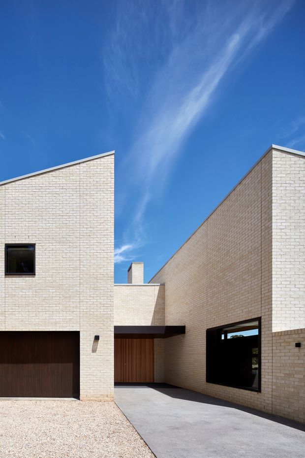 A contemporary residential exterior viewed from the driveway, with intersecting angular brick volumes and crisp sloping rooflines. Pale cream brick walls contrast with dark timber garage and entry doors, black-framed windows, and a charcoal-grey paved driveway. The composition is strongly geometric and minimalist beneath a clear blue sky.