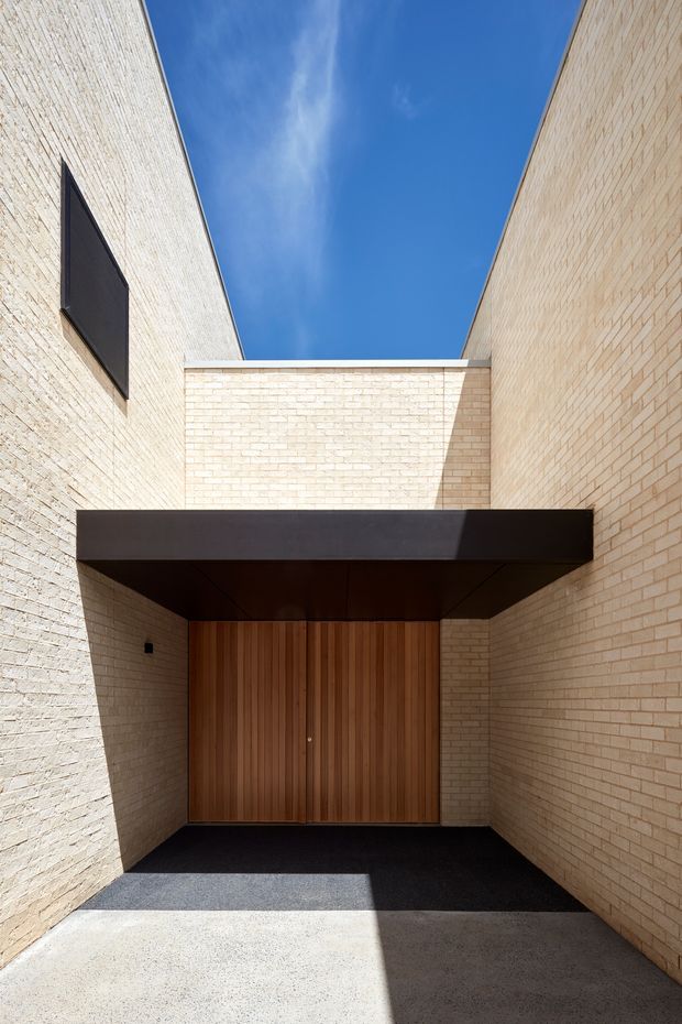 A minimalist architectural exterior showing a recessed entry courtyard framed by tall pale brick walls. A dark projecting canopy shelters a large vertical timber entrance door, with a small dark window or panel high on the left wall. The concrete ground and strong sunlight create pronounced geometric shadows against a clear blue sky.