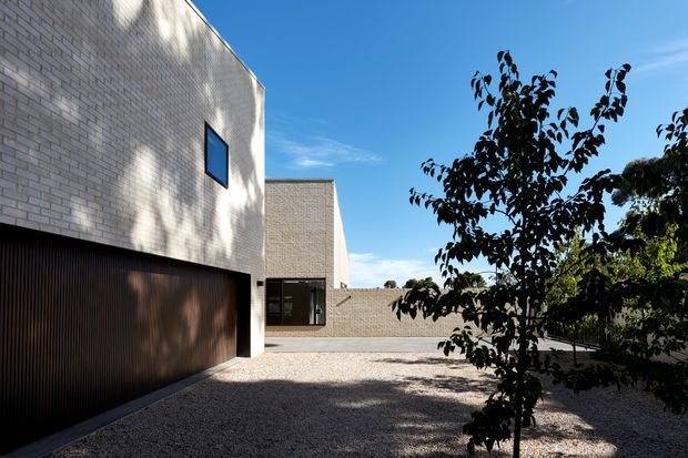 A minimalist contemporary residential exterior viewed from a gravel courtyard or driveway. The building has crisp cubic volumes clad in pale cream brick, a dark vertically ribbed garage door, narrow dark-framed windows, and a low connecting wall. Mature planting casts shadows across the gravel, with strong sunlight and a clear blue sky creating a quiet, geometric composition.
