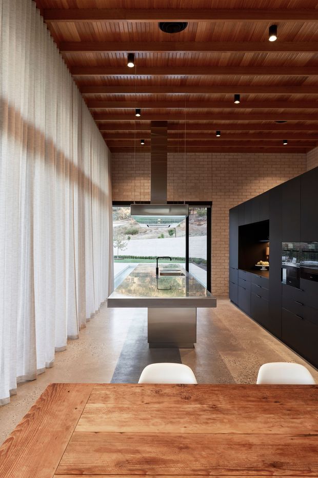 A warm, contemporary kitchen and dining space with a long timber dining table in the foreground, a central island with integrated cooktop and rangehood, dark matte cabinetry along the right wall, exposed light brick walls, and a richly timber-lined ceiling with recessed and track-style spot lighting. Full-height sheer curtains line the left side, while glazing at the rear opens toward an outdoor pool or courtyard.