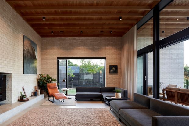 A spacious contemporary living room with pale exposed brick walls, a warm timber-lined ceiling with recessed spotlights, large black-framed glazing, and full-height curtains. The room contains a dark sectional sofa, a rust-orange lounge chair, a pale stone or concrete fireplace hearth, a textured neutral rug, and restrained artwork and greenery. Outdoor dining and landscaped surroundings are visible through the glazing.