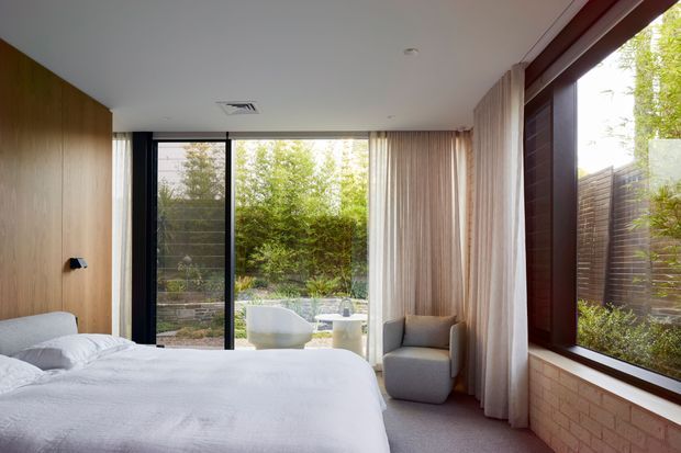 A bright, contemporary bedroom with a low-profile bed, warm timber wall panelling, floor-to-ceiling glazing, and full-height sheer curtains. The room opens visually to a landscaped outdoor area with a freestanding bath visible beyond the glass. A grey upholstered armchair sits beside the window, while a large dark-framed window and exposed brickwork add architectural texture.