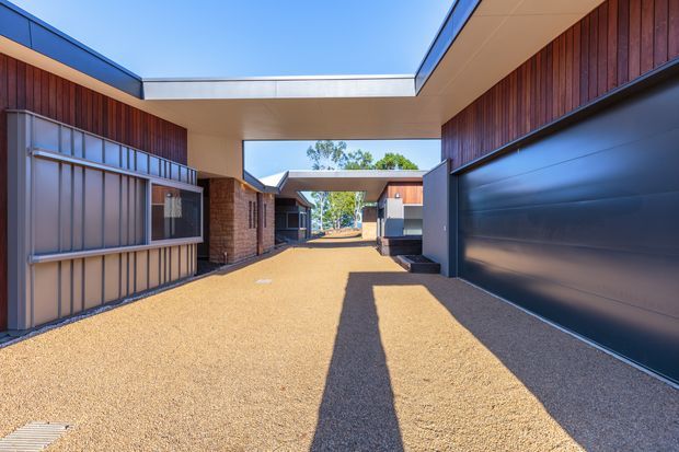 A contemporary residential driveway or courtyard passage framed by connected low-rise structures. The space features dark metal garage or entry doors, warm vertical timber cladding, exposed brick walls, smooth light soffits, and a broad flat-roofed overhead connection. A gravel or fine aggregate surface runs through the open-air approach, with strong sunlight and deep architectural shadows.