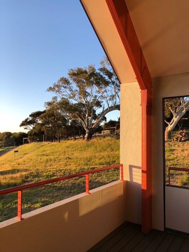 A covered outdoor balcony or veranda overlooking a grassy rural landscape with mature trees. The space has cream rendered walls, dark timber decking, and distinctive red-painted metal railings and structural trims, illuminated by warm late-afternoon sunlight. A doorway or opening at the right leads indoors.