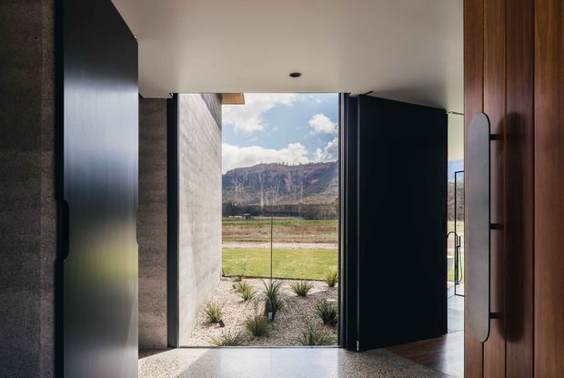 A minimalist modern entryway viewed from inside, with a large open dark-framed door leading to a landscaped front area and expansive mountain landscape beyond. The interior features a smooth light ceiling, polished grey concrete-like floor, dark wall surfaces, and a warm vertical timber door or wall panel with a tall sculptural handle. Strong indoor-outdoor connection and framed mountain views dominate the composition.