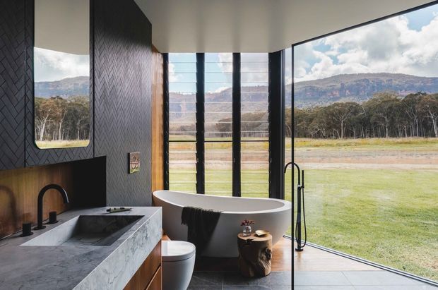 A contemporary bathroom with a freestanding oval bathtub positioned beside full-height glazing overlooking a rural landscape. The room features a dark textured herringbone wall finish, large-format dark floor tiles, a grey stone vanity benchtop with integrated basin, black tapware and shower fittings, timber-toned cabinetry, and a rounded wall mirror. Strong indoor–outdoor connection and abundant natural light define the space.