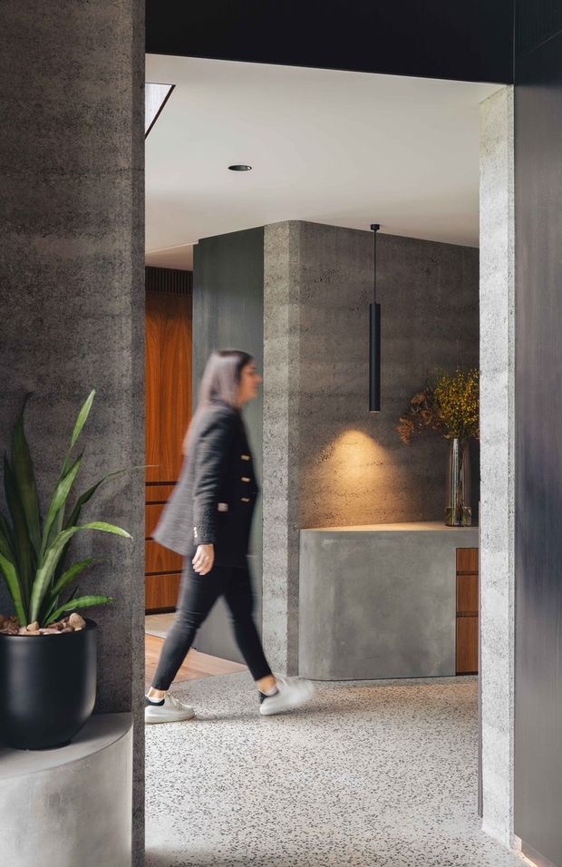 A refined modern entryway featuring dark textured concrete or plaster walls, a polished terrazzo floor, a sculptural black pendant light, and a concrete console with timber drawers. A timber pivot door and indoor plant add warmth, while a person walks through the space.