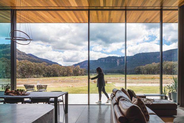 A contemporary open-plan living and dining space with full-height black-framed glazing overlooking a broad rural valley and dramatic mountain escarpment. The room features a warm timber-lined ceiling, polished dark flooring, a large dining table, curved stone island or bench in the foreground, and a substantial brown leather sectional sofa. A person stands near the glazing, while a sculptural linear pendant hangs over the dining area.