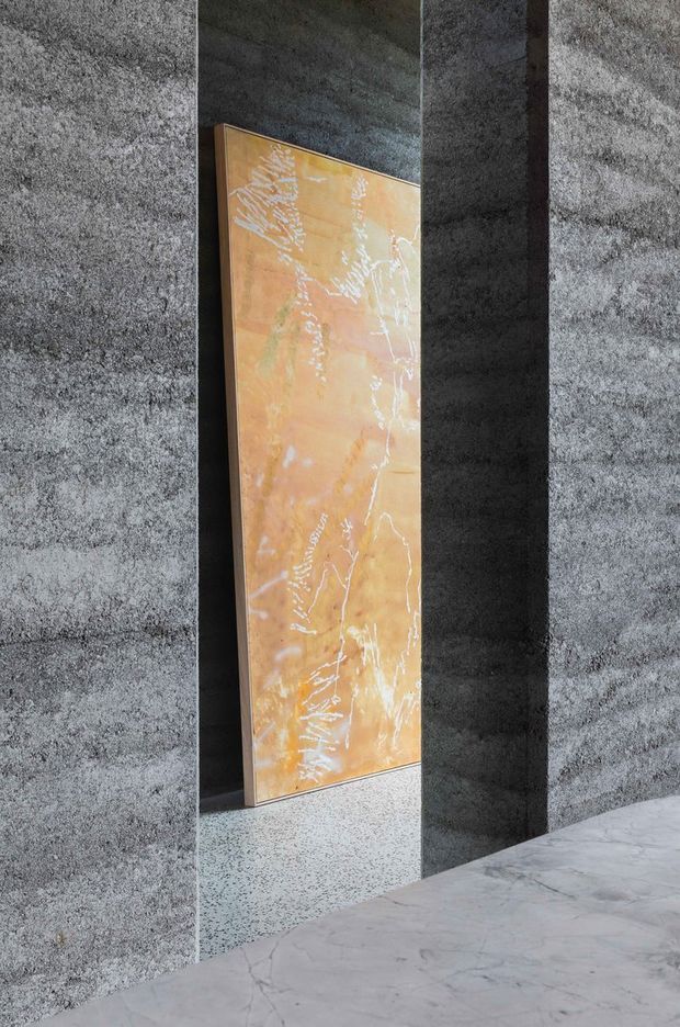 A narrow architectural interior view framed by dark, textured grey stone or concrete piers. A tall amber-orange abstract feature panel with pale organic veining stands in a recessed opening, set against a dark wall. The foreground floor is light grey stone with subtle veining, while a speckled terrazzo-like threshold or floor surface is visible beyond the opening. The composition is minimalist, sculptural, and materially focused.