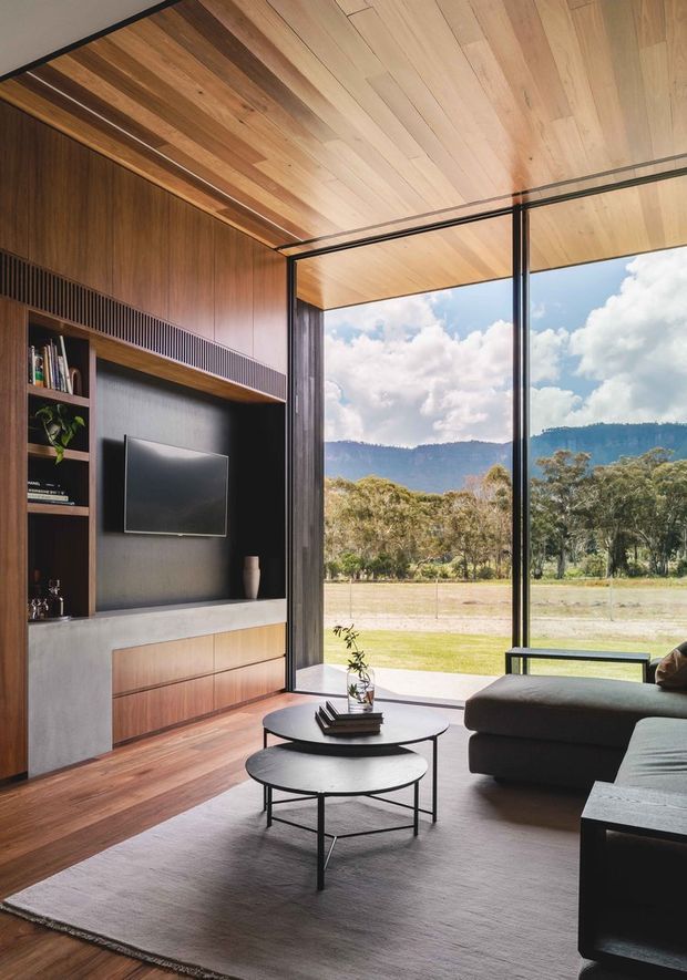 A refined contemporary living room with extensive floor-to-ceiling glazing overlooking a grassy rural landscape and wooded hills. The room features a warm timber-lined ceiling, dark timber built-in media cabinetry with open shelving, a concrete-toned lower cabinet surface, dark sectional seating, nesting black coffee tables, and a large neutral rug. The strong indoor-outdoor connection and exposed timber surfaces create a warm, calm atmosphere.