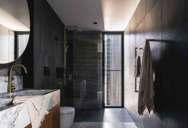 A narrow, sophisticated bathroom with dark large-format wall tiles, a matching dark tiled shower enclosure, and a pale tiled floor. A marble-patterned vanity with timber cabinetry and a curved brass tap occupies the left side, while a wall-mounted brass towel rail and towels line the right wall. A tall narrow window brings in natural light, and a round mirror is partially visible above the vanity.