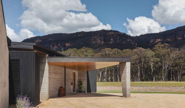 A contemporary single-storey house exterior with a covered carport or entry canopy in the foreground. The structure combines dark vertical cladding, pale rendered or concrete walls, and a warm timber-lined soffit, with the open canopy framing views toward a rural eucalyptus landscape and dramatic sandstone escarpment.