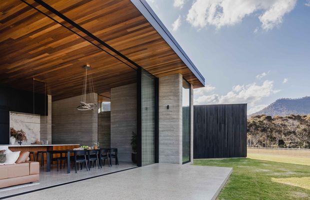 Contemporary single-storey pavilion-style home with a broad covered outdoor terrace opening directly to an open-plan living and dining area. The structure features a warm timber-lined soffit and ceiling, exposed concrete walls and terrace paving, large black-framed glazed sliding doors, and a flat projecting roof. A dining table, kitchen island, and sofa are visible inside, with landscaped lawn and a mountainous backdrop beyond.