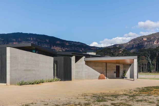 A low, minimalist concrete residence set against a rugged mountain escarpment. The building has broad smooth concrete walls, dark vertical timber or metal-clad entry elements, flat roof planes, and a deep sheltered outdoor entry or terrace with a warm timber-lined soffit. The sparse site and strong horizontal composition emphasise the contemporary architectural form.