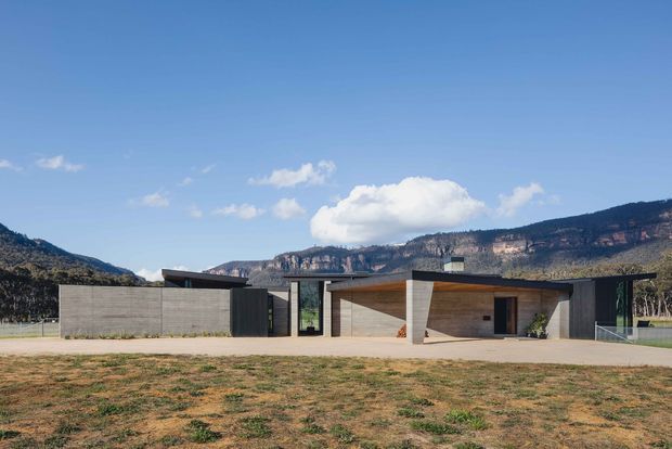 A low, single-storey contemporary residence set against a dramatic mountain landscape. The building has a strongly horizontal composition, flat rooflines, concrete-toned walls, dark vertical cladding or screens, broad glazing, and warm timber lining beneath the entry and carport-like canopy. A paved forecourt and sparse grass occupy the foreground.