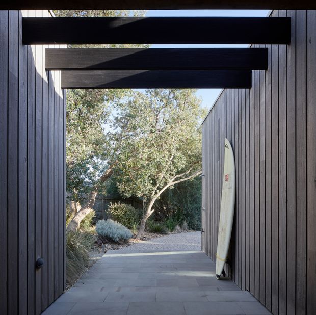 A covered passageway framed by dark vertical timber cladding and heavy exposed overhead beams, opening onto a landscaped garden. Large-format grey paving leads through the shaded entry, with a surfboard stored against the wall and native-style planting visible beyond.