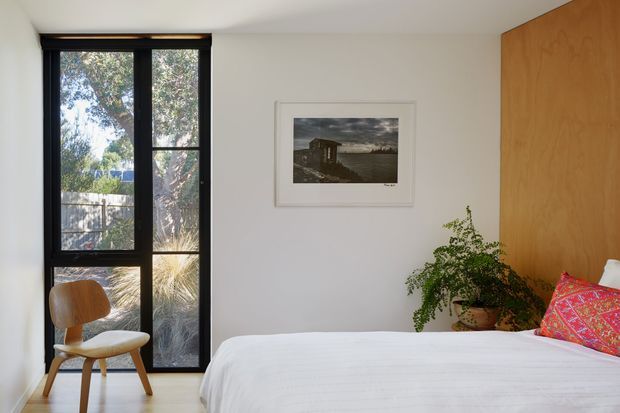 A calm, minimalist bedroom with a low-profile bed dressed in white linen, a warm timber feature wall and matching timber door, black-framed full-height window, and a simple wooden chair. A potted plant and framed artwork add understated character. Natural light creates a bright, quiet atmosphere.