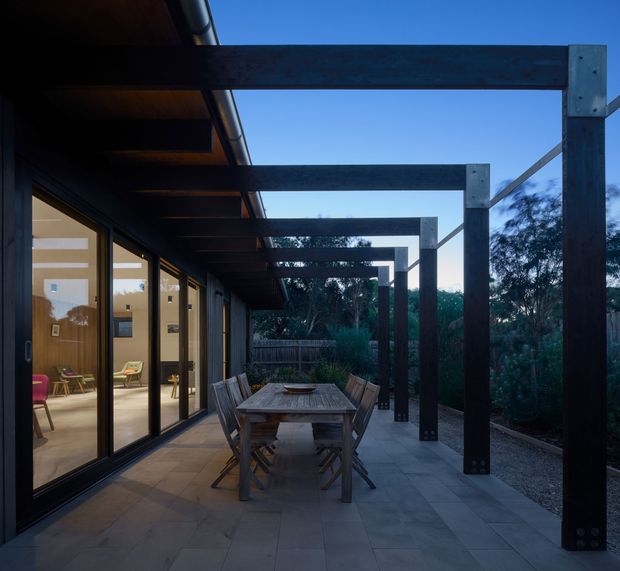 A covered outdoor terrace beside a glazed living area, featuring a large timber dining table and folding chairs on grey tiled paving. A dark timber or steel pergola structure extends overhead and frames the edge of the terrace, with landscaped planting and fencing beyond. Warm interior lighting is visible through the full-height sliding glass doors, creating a calm transition between indoors and outdoors.