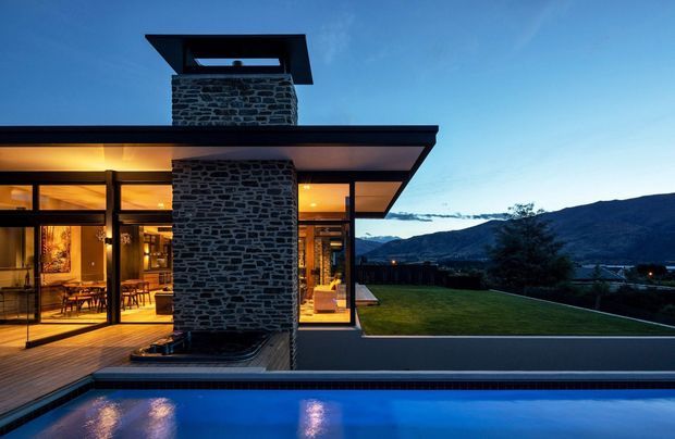 Twilight exterior view of a contemporary, single-storey home beside a swimming pool. The building has extensive floor-to-ceiling glazing, a broad flat roof with deep overhangs, and a prominent central chimney clad in dark irregular stone. Warm interior lighting reveals an open dining and living area, while a manicured lawn and mountainous landscape extend beyond the house.