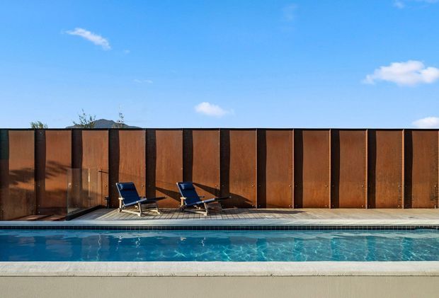 A modern outdoor swimming pool bordered by a narrow tiled deck and two lounge chairs. A tall, continuous rust-brown metal privacy screen with dark vertical structural divisions forms the backdrop, with a clear blue sky above.