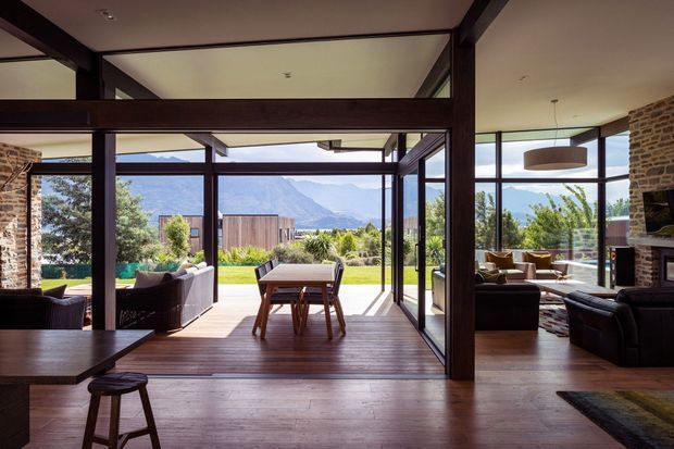 A spacious open-plan interior with dark timber structural beams and framing, exposed brick walls, and extensive floor-to-ceiling glazing opening onto a landscaped outdoor area. The central timber dining table sits between a lounge area and the garden, with mountain views visible beyond. A second seating area and fireplace are visible to the right. The room has a warm, contemporary lodge-like character and strong indoor-outdoor connection.